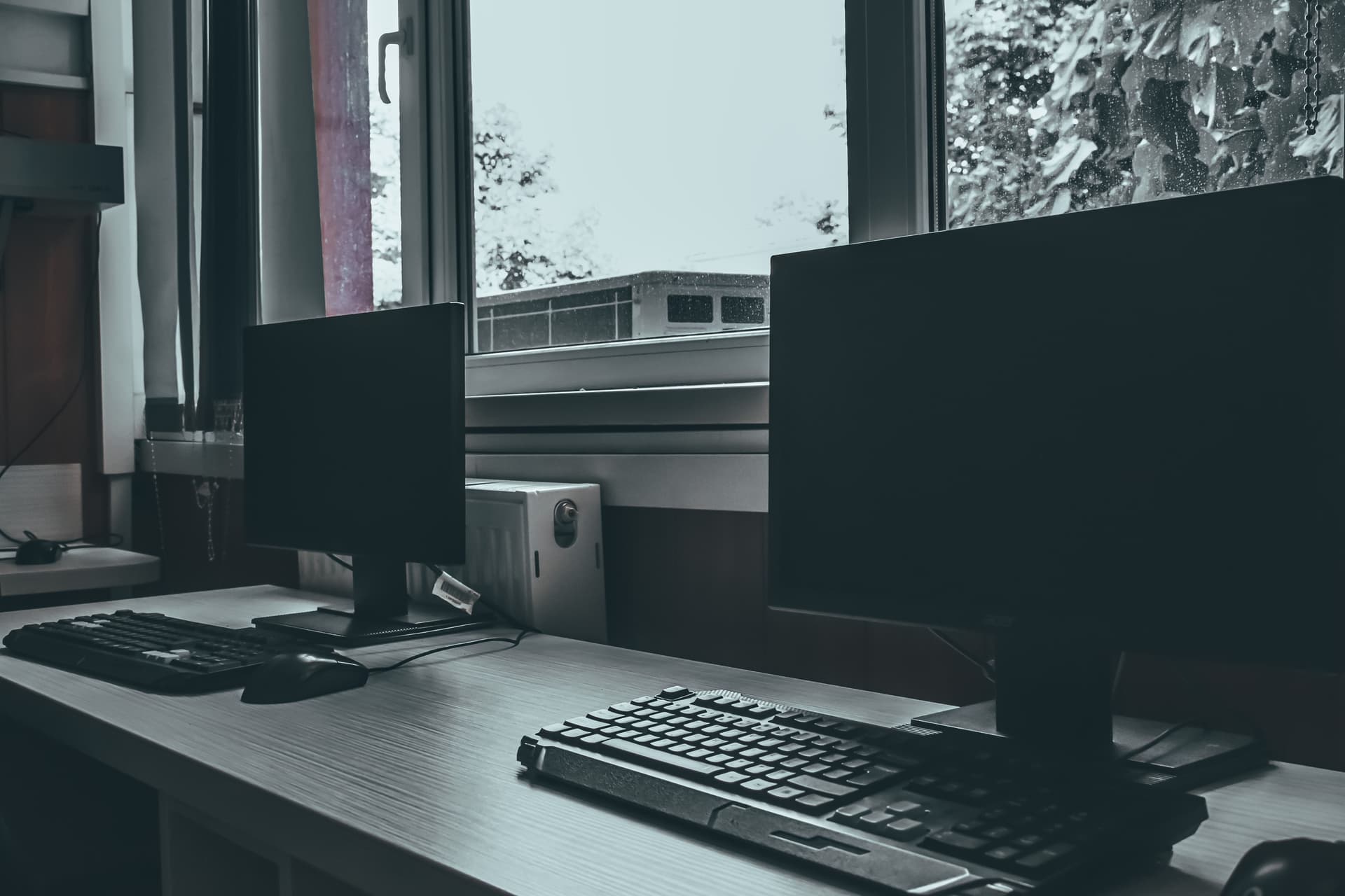 An empty desk by a window with two dark monitors and two keyboards on it.
